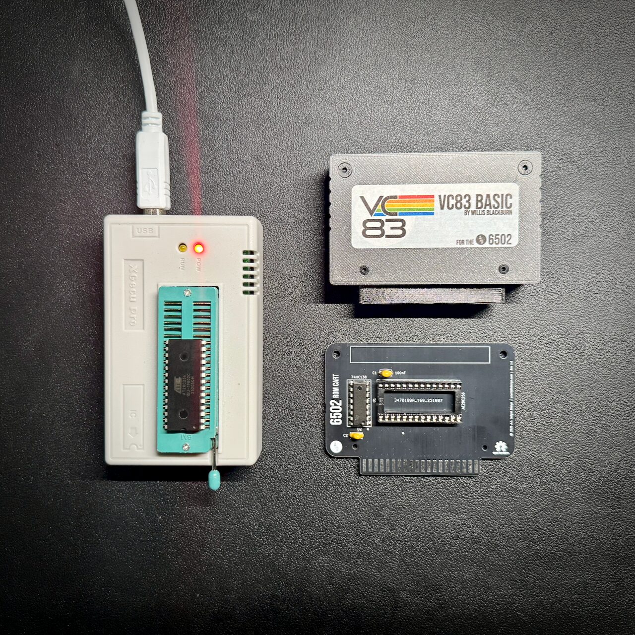 A chip programmer with an EEPROM seated in its ZIF socket and a red LED lit, next to a finished cartridge board and its printed VC83 BASIC label.