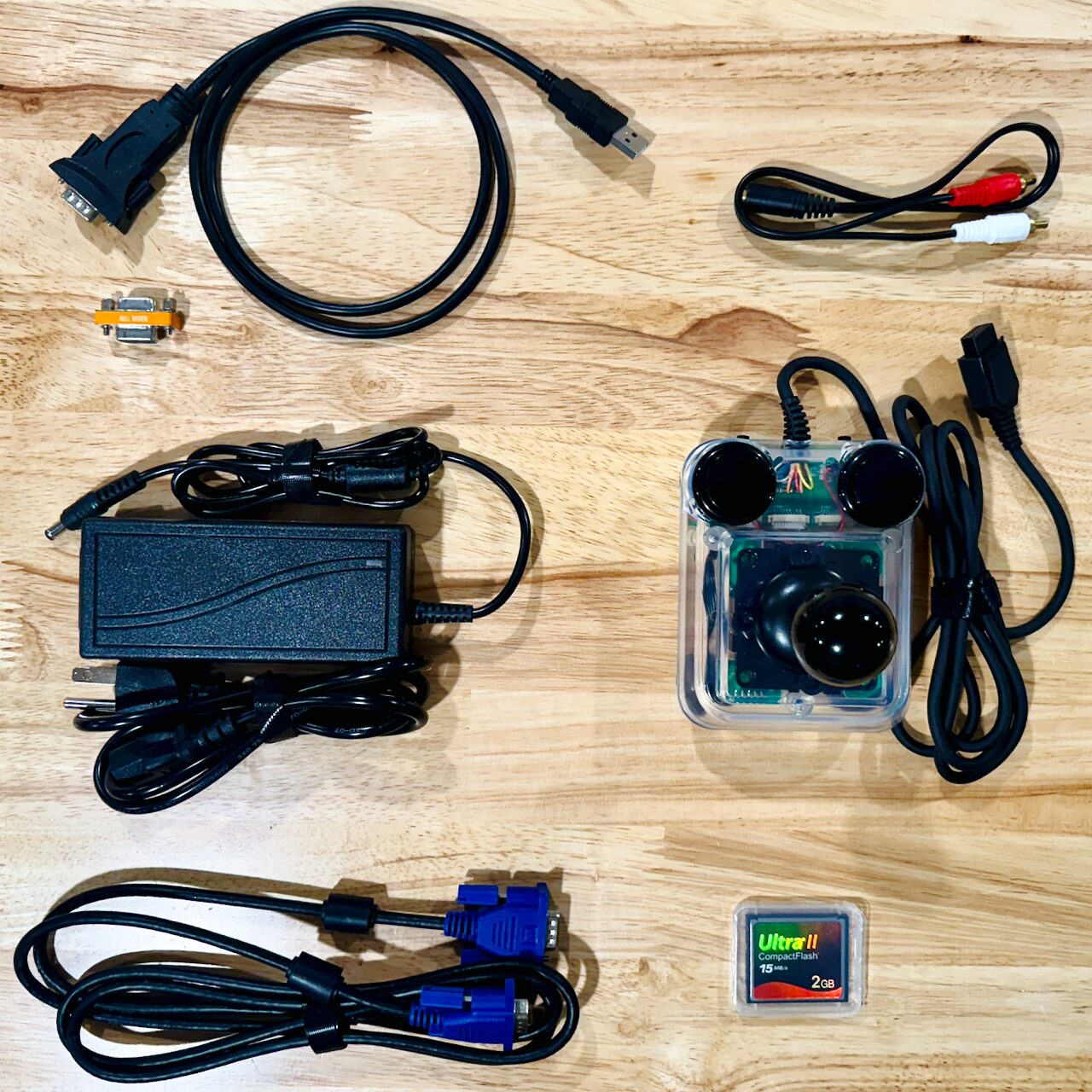 A VGA cable, an audio cable, a power supply, a serial null-modem adapter, a joystick, and a CompactFlash card, laid out on a wooden table.