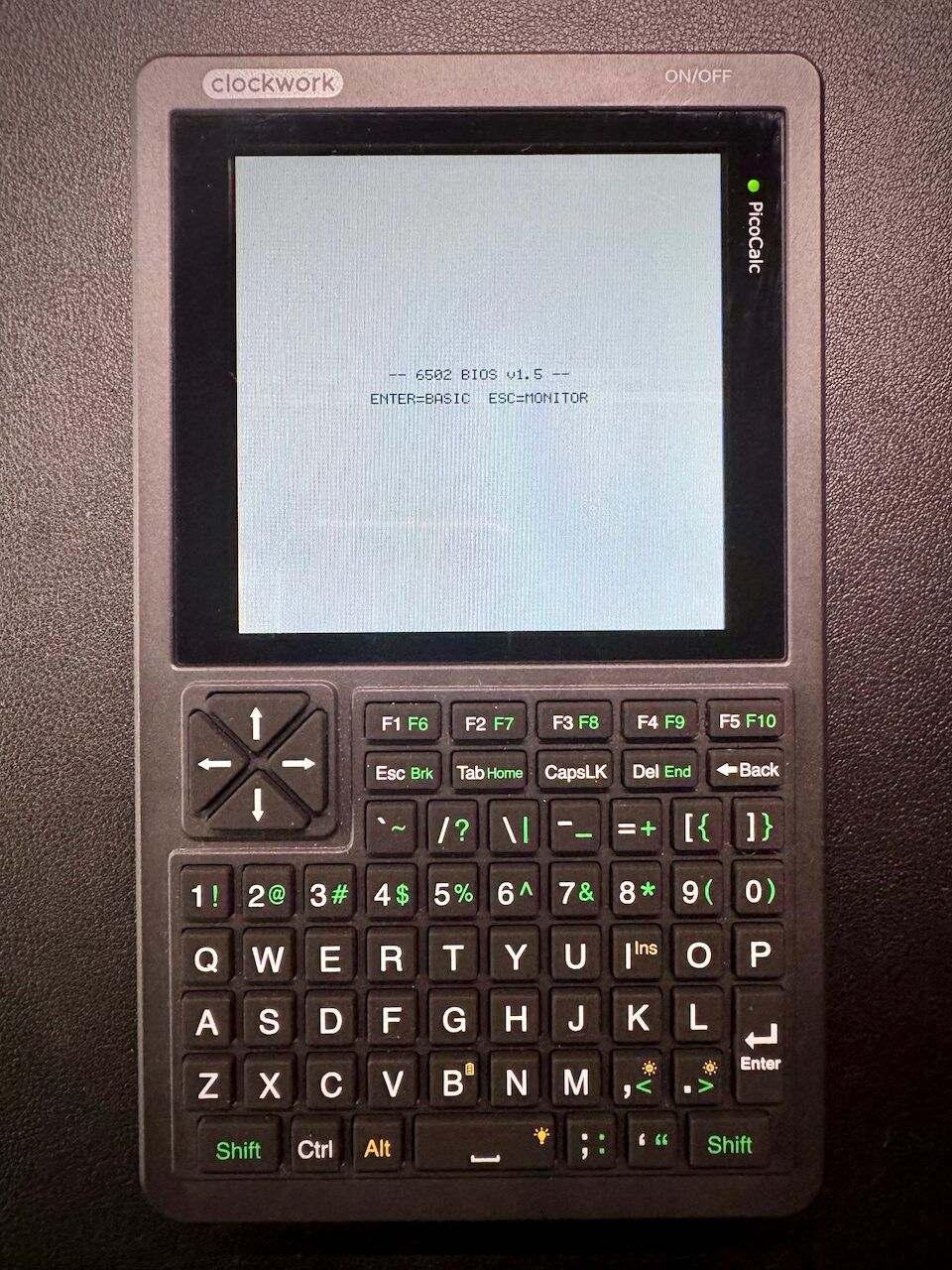 A PicoCalc handheld: a square LCD showing the BIOS splash and its two-line menu, above a QWERTY keyboard with a four-way pad.